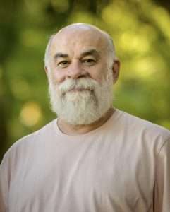 Homem de barba branca e camiseta clara sorri serenamente em ambiente ao ar livre com fundo desfocado de árvores.