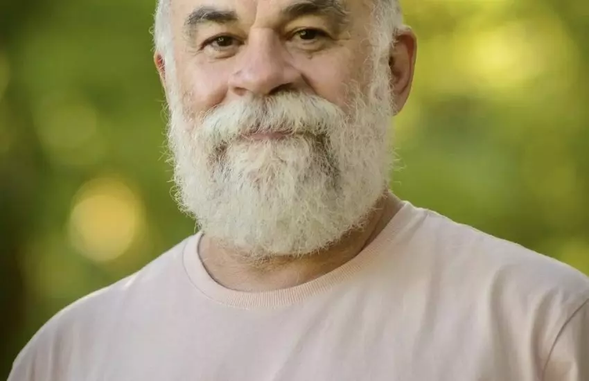 Homem de barba branca e camiseta clara sorri serenamente em ambiente ao ar livre com fundo desfocado de árvores.